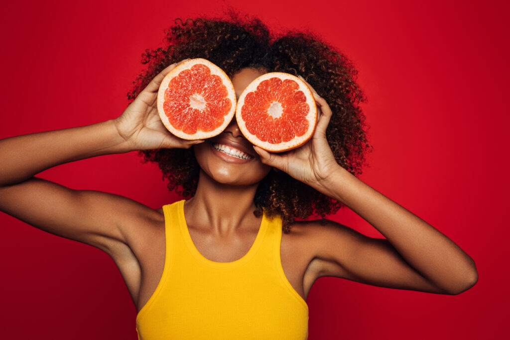 smiling woman in yellow tank top holding two grapefruit halves in front of her eyes