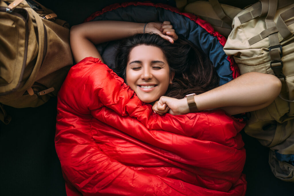 The Traveler Is Resting In A Sleeping Bag. Rest Concept. A Tourist Is Resting In A Tent. Woman Relaxing In Sleeping Bag. Travel, Camping Concept, Adventure. Happy Smiling Female Traveler. The Traveler