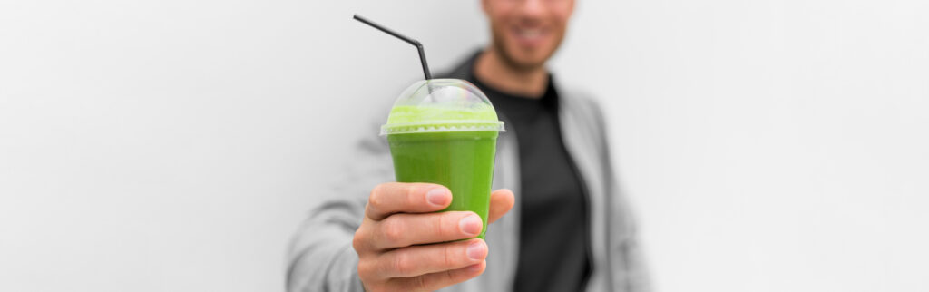 Green Smoothie Juice Drink At Cafe Banner. Healthy Man Drinking Detox Diet Protein Shake White Background Panoramic. Hand Holding Plastic Cup Of Vegetable Juice, Health Food Eating Lifestyle.