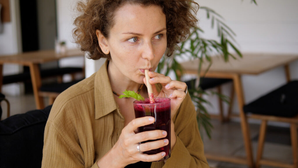 A Young Caucasian Woman Enjoys Her Healthy Organic Cold Pressed Beetroot Juice. Sipping It Through The Paper Straw In A Tropical Cafe.useful Detox Food From Fresh Fruits.